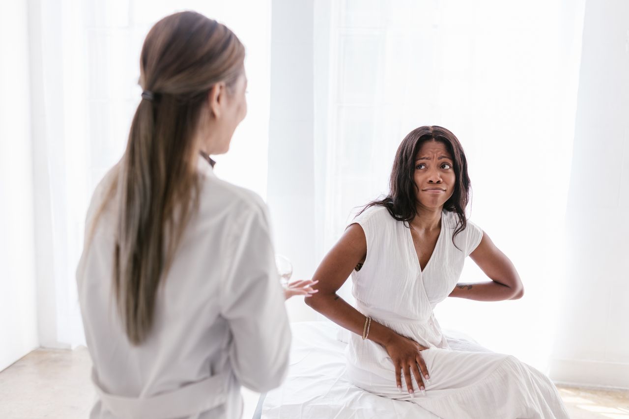 A woman with back pain consults her doctor in a clinical setting wearing a white dress.